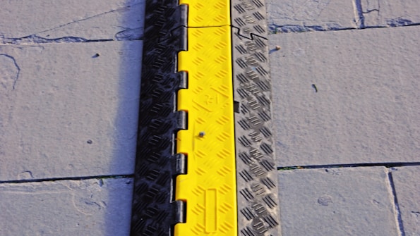 A bright yellow and black cable protector placed on a gray pavement with visible concrete slabs and seams.