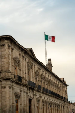A grand historical building with elaborate stone architecture and numerous balconies. A large flagpole hoists the Mexican flag proudly above the structure against a slightly cloudy sky.