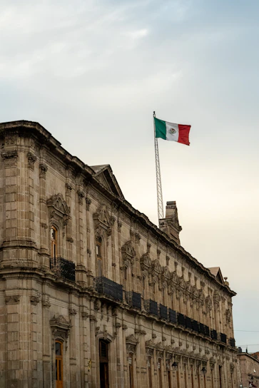 A grand historical building with elaborate stone architecture and numerous balconies. A large flagpole hoists the Mexican flag proudly above the structure against a slightly cloudy sky.