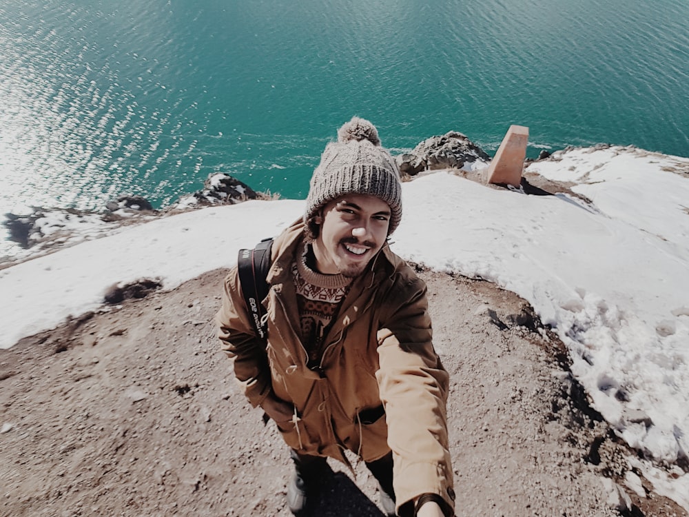 woman in brown jacket and gray knit cap standing on brown rock near body of water