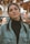 A woman with braided hair stands in an indoor setting wearing a denim jacket over a black turtleneck. She has large hoop earrings and maintains a neutral expression. The background appears to be out-of-focus lights and colors, suggesting a festive or lively atmosphere.
