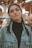 A woman with braided hair stands in an indoor setting wearing a denim jacket over a black turtleneck. She has large hoop earrings and maintains a neutral expression. The background appears to be out-of-focus lights and colors, suggesting a festive or lively atmosphere.