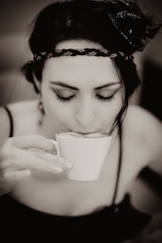 A woman is sipping from a small white cup, eyes closed, wearing a decorative headband with detailed braiding. The image is captured in a soft sepia tone, giving it a vintage feel.