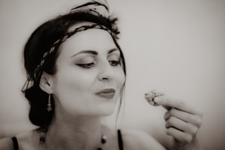 A woman with dark hair tied back and wearing a decorative headband and earrings is looking at a small item held in her hand, possibly food or a trinket. Her makeup is done in a vintage style, and the image has a sepia tone, giving it a nostalgic feel.