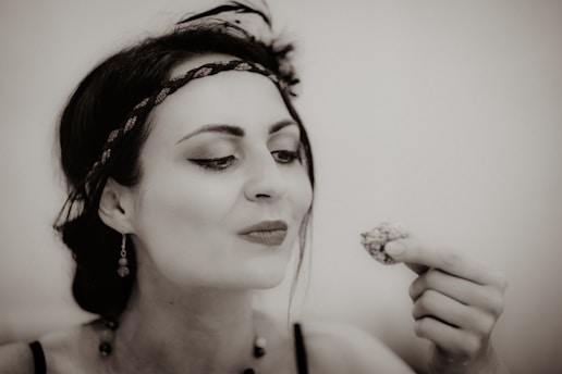 A woman with dark hair tied back and wearing a decorative headband and earrings is looking at a small item held in her hand, possibly food or a trinket. Her makeup is done in a vintage style, and the image has a sepia tone, giving it a nostalgic feel.