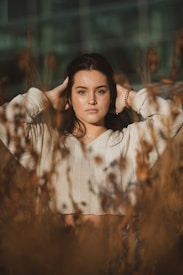 A person stands in a field with tall dry grasses, wearing a cream-colored sweater with arms raised behind the head. The expression is calm and introspective.