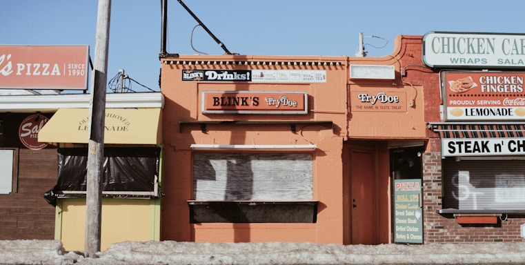 A row of small, rustic restaurants and shops with colorful signs, including a pizza place, lemonade stand, and chicken cafe. The buildings have a vintage aesthetic with signs advertising various foods like steak, cheese, and drinks.