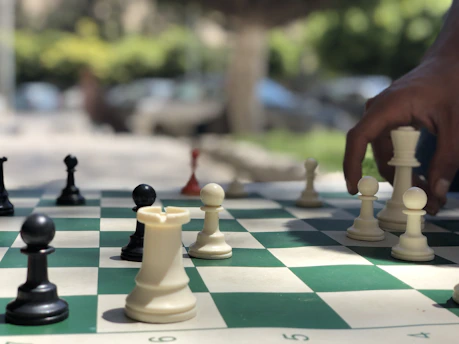 A chess game in progress outdoors, with a hand reaching to move a white piece. The board features both black and white chess pieces in various positions, implying a strategic moment in the game. The background is blurred, showing trees and a few vehicles.