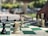 A chess game in progress outdoors, with a hand reaching to move a white piece. The board features both black and white chess pieces in various positions, implying a strategic moment in the game. The background is blurred, showing trees and a few vehicles.
