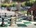 A chess game in progress outdoors, with a hand reaching to move a white piece. The board features both black and white chess pieces in various positions, implying a strategic moment in the game. The background is blurred, showing trees and a few vehicles.
