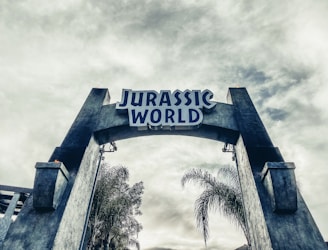 A large stone archway with the words 'Jurassic World' prominently displayed at the top. The structure is flanked by tall, weathered stone pillars. In the background, there are palm trees and a cloudy sky, adding a prehistoric or adventurous ambiance.