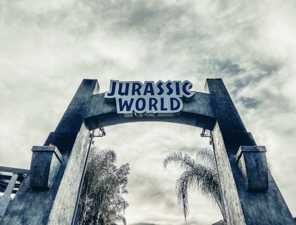 A large stone archway with the words 'Jurassic World' prominently displayed at the top. The structure is flanked by tall, weathered stone pillars. In the background, there are palm trees and a cloudy sky, adding a prehistoric or adventurous ambiance.