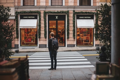 A person stands facing a luxury store, specifically Louis Vuitton, across a crosswalk. The store's facade is adorned with green ivy, and the display windows showcase various high-end products. The scene suggests an urban setting with a touch of sophistication.