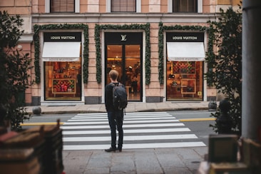 A person stands facing a luxury store, specifically Louis Vuitton, across a crosswalk. The store's facade is adorned with green ivy, and the display windows showcase various high-end products. The scene suggests an urban setting with a touch of sophistication.