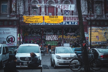 A street scene features a building facade covered in colorful graffiti, with prominent posters and banners protesting political issues. Several cars are parked in front, and a few people and bicycles are scattered around the area. The banners and graffiti convey a sense of activism and urban culture.