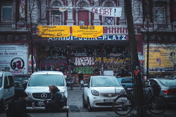 A street scene features a building facade covered in colorful graffiti, with prominent posters and banners protesting political issues. Several cars are parked in front, and a few people and bicycles are scattered around the area. The banners and graffiti convey a sense of activism and urban culture.