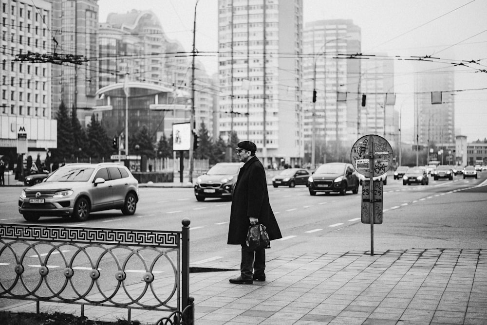 grayscale photo of man in black coat standing near road