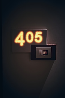 A dimly lit hotel room number sign displays the number 405. Next to it, there is a small control panel featuring a bell icon and a do-not-disturb symbol, which is illuminated. The shadows cast on the textured wall add an atmospheric touch.