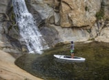 A person stands on a paddleboard in a small pool of water near a rocky waterfall. The individual is holding a paddle and appears to be enjoying a tranquil moment surrounded by natural rock formations.