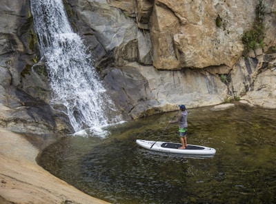 A person stands on a paddleboard in a small pool of water near a rocky waterfall. The individual is holding a paddle and appears to be enjoying a tranquil moment surrounded by natural rock formations.
