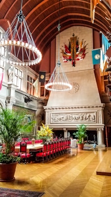 An ornate, grand room with a large fireplace adorned with intricate carvings. The space features a high, arched wooden ceiling and two large chandeliers with lit bulbs. A long dining table is set with red chairs, and the room is decorated with indoor plants and vibrant flower arrangements. Tapestries and flags hang from the upper walls, adding to the luxurious feel.
