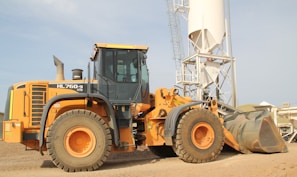 A large yellow construction vehicle, specifically a front loader, is situated on a gravel-covered ground. Behind it, a tall white industrial silo with a metal frame stands prominently against a clear sky. The loader is equipped with large tires and a sizable bucket. The environment appears industrial, with additional structures visible in the background.