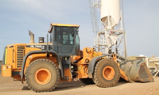 A large yellow construction vehicle, specifically a front loader, is situated on a gravel-covered ground. Behind it, a tall white industrial silo with a metal frame stands prominently against a clear sky. The loader is equipped with large tires and a sizable bucket. The environment appears industrial, with additional structures visible in the background.