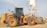A large yellow construction vehicle, specifically a front loader, is situated on a gravel-covered ground. Behind it, a tall white industrial silo with a metal frame stands prominently against a clear sky. The loader is equipped with large tires and a sizable bucket. The environment appears industrial, with additional structures visible in the background.