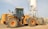 A large yellow construction vehicle, specifically a front loader, is situated on a gravel-covered ground. Behind it, a tall white industrial silo with a metal frame stands prominently against a clear sky. The loader is equipped with large tires and a sizable bucket. The environment appears industrial, with additional structures visible in the background.