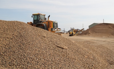 A large industrial vehicle parked beside a substantial pile of gravel, with additional mounds of gravel and industrial structures in the background. The scene appears to be a gravel or construction site under clear skies.