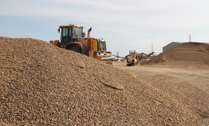 A large industrial vehicle parked beside a substantial pile of gravel, with additional mounds of gravel and industrial structures in the background. The scene appears to be a gravel or construction site under clear skies.