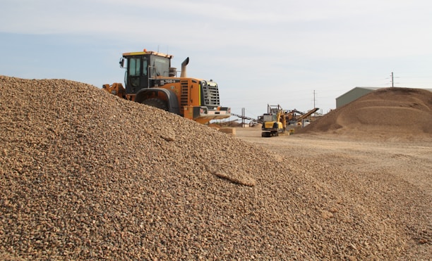 A large industrial vehicle parked beside a substantial pile of gravel, with additional mounds of gravel and industrial structures in the background. The scene appears to be a gravel or construction site under clear skies.