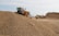 A large industrial vehicle parked beside a substantial pile of gravel, with additional mounds of gravel and industrial structures in the background. The scene appears to be a gravel or construction site under clear skies.