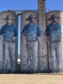 Three large murals painted on concrete silos depict a man wearing a wide-brimmed hat and a blue shirt. Each figure holds a long branch, and the murals show the man in different poses, looking in various directions. The artwork is detailed, showcasing realistic features against a clear blue sky backdrop.