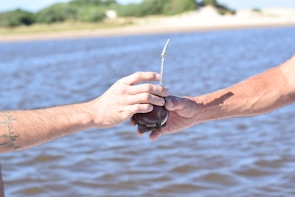Two hands extend toward each other, one offering a traditional mate cup with a metal straw, against the backdrop of a rippling body of water and distant greenery.