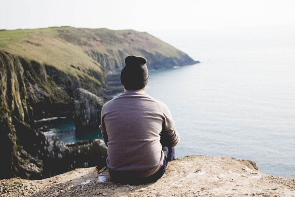 man in gray hoodie sitting on brown rock near body of water during daytime