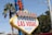 A retro-style sign with large, bold letters welcomes visitors to Las Vegas. The sign features neon elements and vibrant colors, framed by palm trees and electrical lines against a clear blue sky.