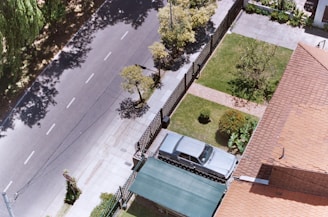 An aerial view of a residential neighborhood displays a quiet street lined with trees casting shadows. A driveway leads to a garage with a parked silver car. The adjacent house, featuring a red-tiled roof, has a neatly maintained garden with greenery and small trees.