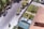 An aerial view of a residential neighborhood displays a quiet street lined with trees casting shadows. A driveway leads to a garage with a parked silver car. The adjacent house, featuring a red-tiled roof, has a neatly maintained garden with greenery and small trees.