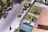An aerial view of a residential neighborhood displays a quiet street lined with trees casting shadows. A driveway leads to a garage with a parked silver car. The adjacent house, featuring a red-tiled roof, has a neatly maintained garden with greenery and small trees.