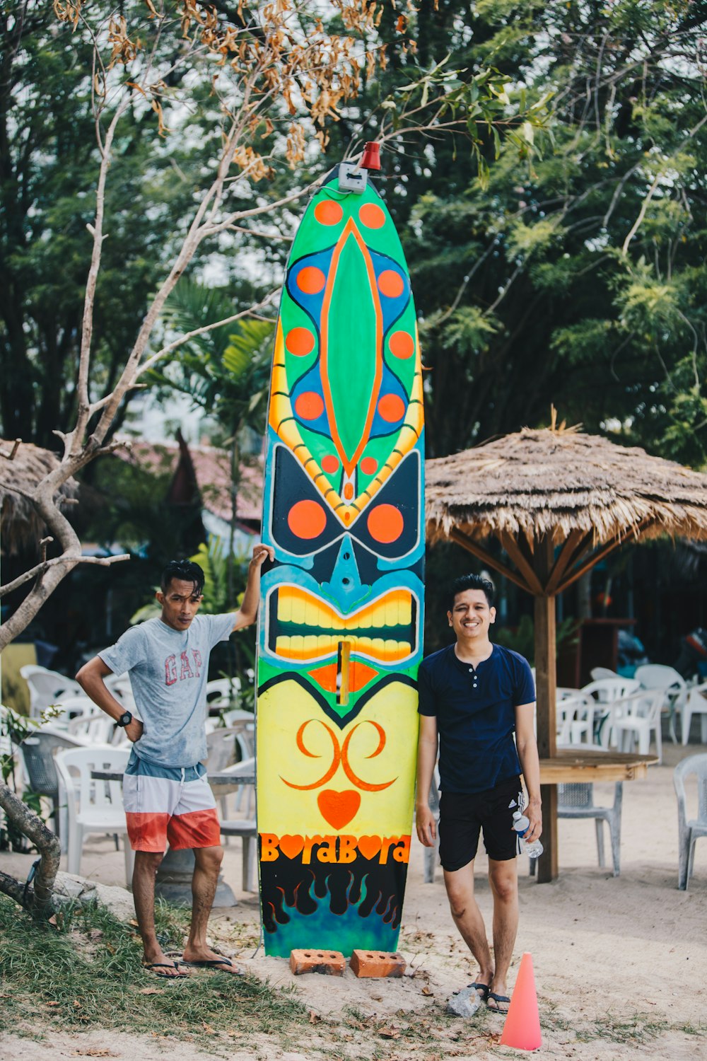 Hombre con camiseta negra de cuello redondo de pie junto a una tabla de surf azul y amarilla