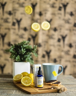Several slices of lemon are suspended in mid-air in front of a rustic background. On a wooden surface, a small cutting board displays a few whole and halved lemons, a bottle of CBD oil with its box, a cup with a tea bag tag hanging out, and a couple of chocolate-topped biscuits. A potted plant adds some greenery to the composition.
