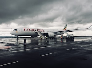 A large passenger airplane parked on a wet runway under a cloudy sky. The airplane is branded with 'Ethiopian' on the side, and there is a staircase leading up to it. Ground crew members are visible around the aircraft, some wearing reflective vests.