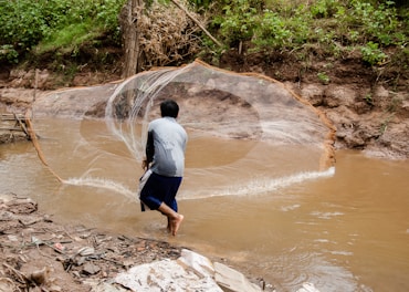 A person is standing knee-deep in a muddy river, casting a large fishing net. The surroundings are natural with green foliage and a muddy riverbank. The water appears brown due to sediment, and the person is wearing a long-sleeved shirt and shorts.