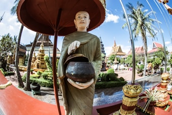 A statue of a monk holding a bowl stands prominently in a garden filled with palm trees and traditional temple structures. The scene is bright and colorful, with architectural elements suggesting a Southeast Asian temple setting. The blue sky above contrasts with the intricate gold and brown tones of the temple buildings.