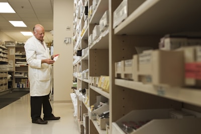 A person in a white lab coat is standing in front of a series of shelves filled with various boxed items, likely in a pharmacy or medical supply room. The individual is focused on examining a box held in their hand, suggesting they are organizing or checking inventory.