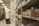 A person in a white lab coat is standing in front of a series of shelves filled with various boxed items, likely in a pharmacy or medical supply room. The individual is focused on examining a box held in their hand, suggesting they are organizing or checking inventory.