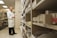 A person in a white lab coat is standing in front of a series of shelves filled with various boxed items, likely in a pharmacy or medical supply room. The individual is focused on examining a box held in their hand, suggesting they are organizing or checking inventory.