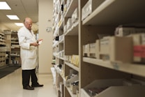 A person in a white lab coat is standing in front of a series of shelves filled with various boxed items, likely in a pharmacy or medical supply room. The individual is focused on examining a box held in their hand, suggesting they are organizing or checking inventory.