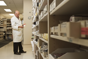 A person in a white lab coat is standing in front of a series of shelves filled with various boxed items, likely in a pharmacy or medical supply room. The individual is focused on examining a box held in their hand, suggesting they are organizing or checking inventory.